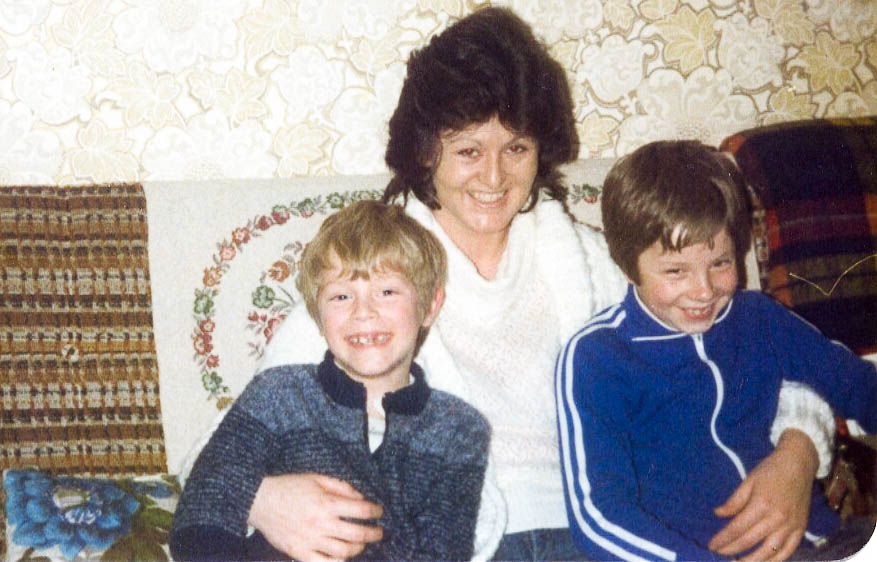 Two brothers aged around 8 and 10 with their older female cousin in the middle, all smiling and sitting on a sofa with a real 1980s pattern on the sofa and on the wallpaper behind.